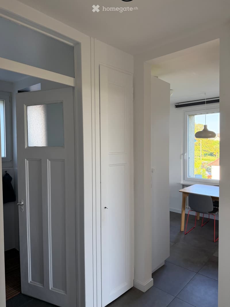 A modern hallway with white walls and doors leading to different rooms. A small table and chair are visible in the adjacent room with a window showing a view of greenery.