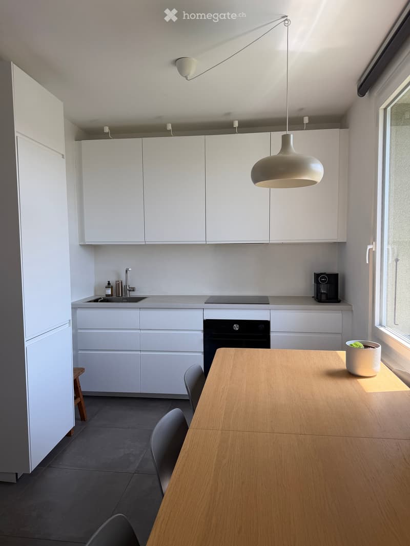 Modern kitchen with white cabinets, a wooden dining table, grey chairs, and a pendant light. Features a sink, faucet, oven, and coffee maker on the counter.