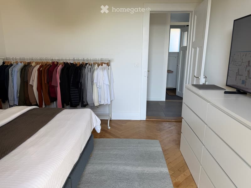 Modern bedroom with a neatly made bed, a white dresser with a TV on top, a clothing rack with various garments, and a grey rug on the wooden floor.