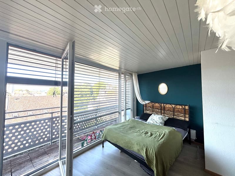 Bedroom with a large bed, decorative headboard, pillows, and a mirror on the wall. Large windows open to a balcony with a view of rooftops.