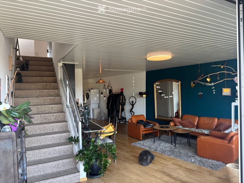 Modern living room with wooden flooring, dark blue accent wall, leather sofa, small tables, decorative mirror, and staircase leading to another level.
