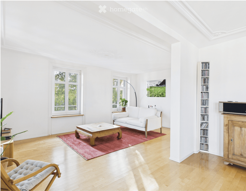 Bright living room with white walls, wooden flooring, and large windows. Features a white sofa, wooden coffee table, armchair, bookshelf, floor lamp, and a potted plant.