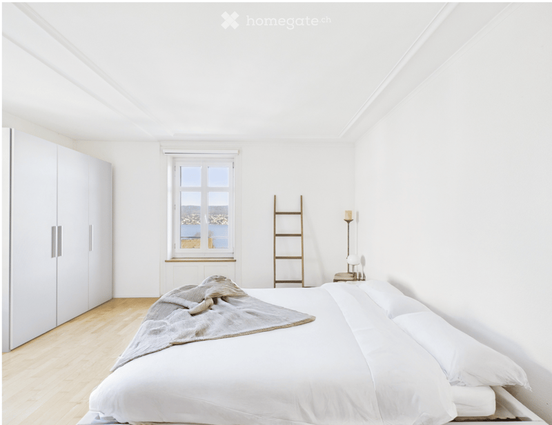 Bright, minimalist bedroom with white walls, wooden floor, large window offering a scenic view, bed with white linens, nightstand with lamp, wardrobe, and a wooden ladder leaning against the wall.