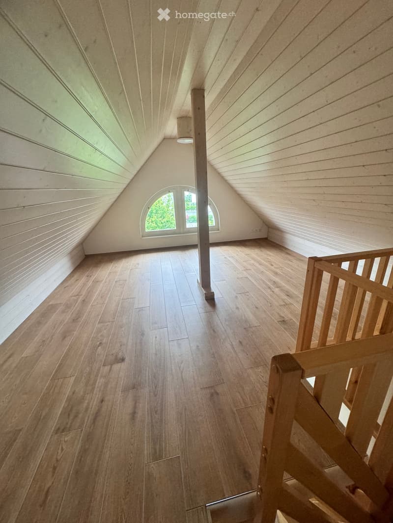 Empty attic space with wooden flooring, slanted wooden ceiling, and a small window letting in natural light.