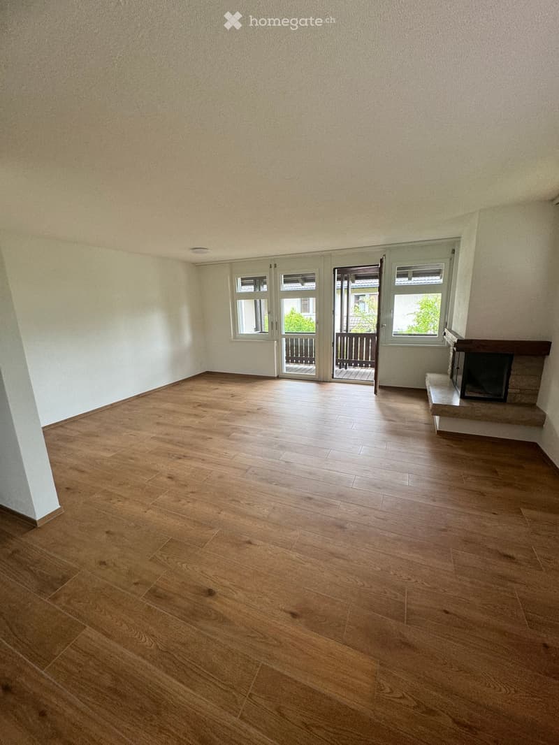 Empty room with wooden flooring, white walls, and a fireplace on the right side. There are large windows and a door leading to a balcony.