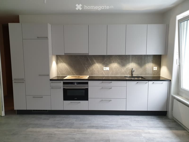 Modern kitchen with white cabinets, stainless steel sink, and built-in oven. Features a sleek, minimalist design with a dark countertop and light-colored floor.
