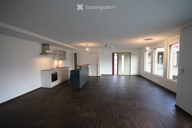 Open-plan space with a modern kitchen area featuring white cabinets, a built-in oven, and a countertop, connected to a living area with large windows and wooden flooring.