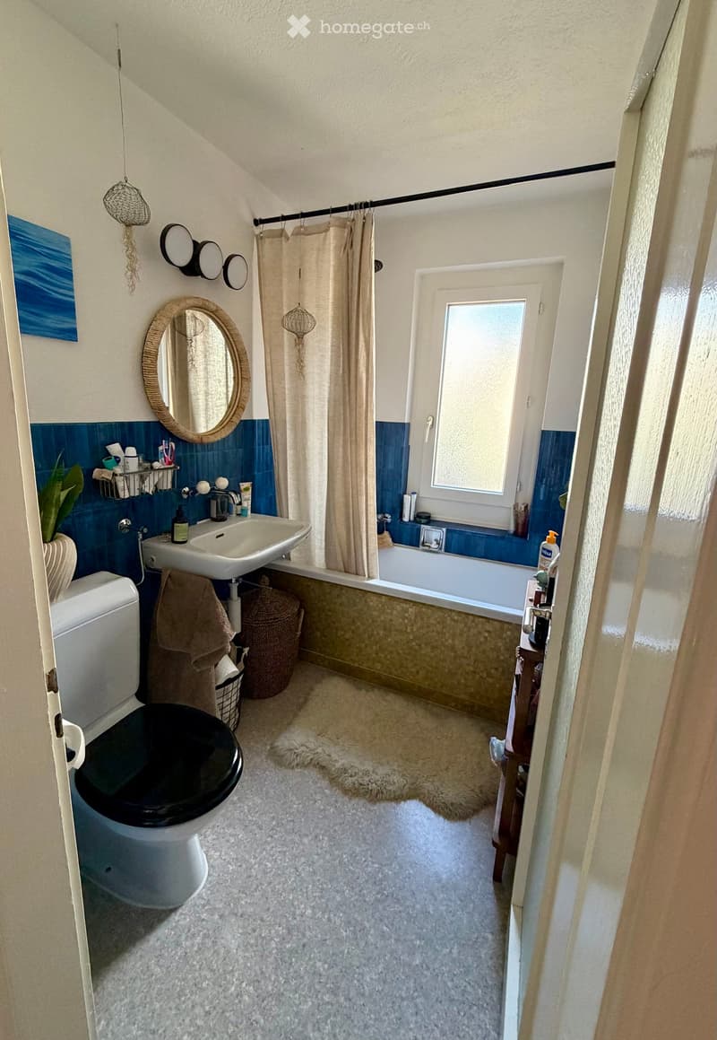 Bathroom with white walls, blue tiles around the sink and bathtub, a large mirror, a shower curtain, a sink, a toilet, a bathtub, a wicker basket, a towel, and a small plant on the shelf.