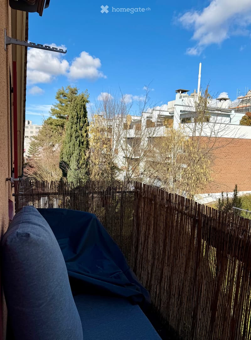 Balcony with a wooden fence, covered seating area, and view of surrounding buildings and trees.