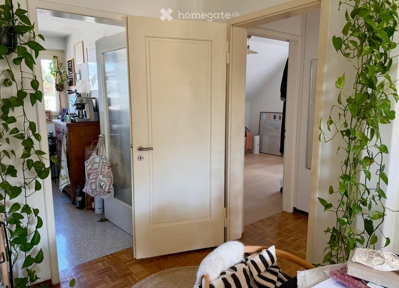 A cozy, well-lit room with wooden flooring, a large window, and a view of the hallway. The room features a comfortable chair with a white fur throw and black-and-white striped cushions, a small table with a plant, and a coffee machine on a wooden cabinet