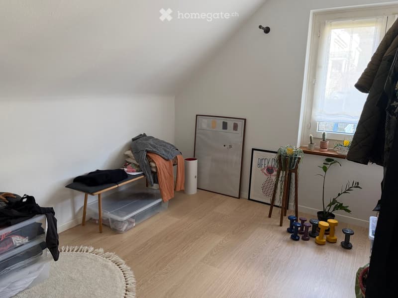 A small, well-lit room with wooden flooring, a bench with folded clothes, storage bins, picture frames, potted plants, and exercise weights.