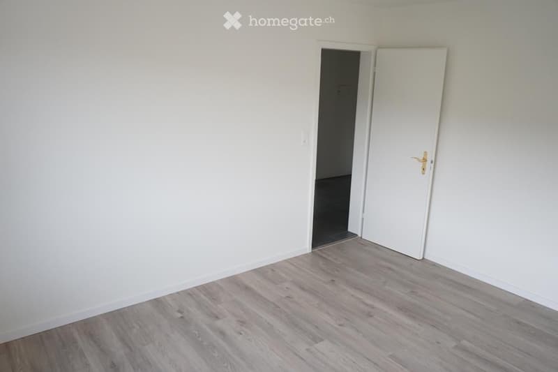 Empty room with light-colored wooden flooring and white walls, featuring two doors, one leading to another room.