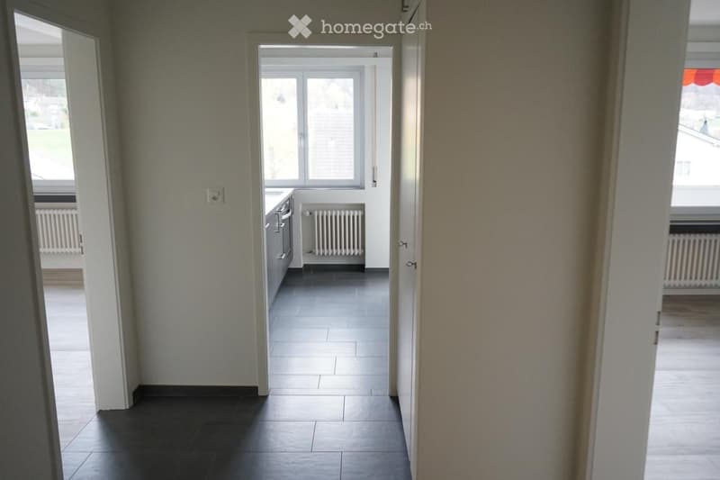 Empty hallway leading to a kitchen area with tiled floors, white walls, and built-in cabinets.
