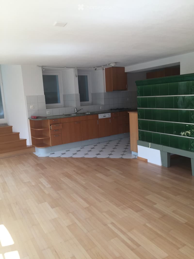 Open-plan kitchen and living area with wooden flooring, wooden cabinets, a sink, and a dishwasher. Green tiled backsplash and a staircase leading upwards.