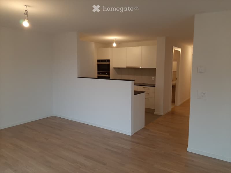 Empty room with wooden flooring, white walls, and a white kitchen counter. The kitchen area includes cabinets, an oven, and a stove, but no furniture or appliances are present in the main space.