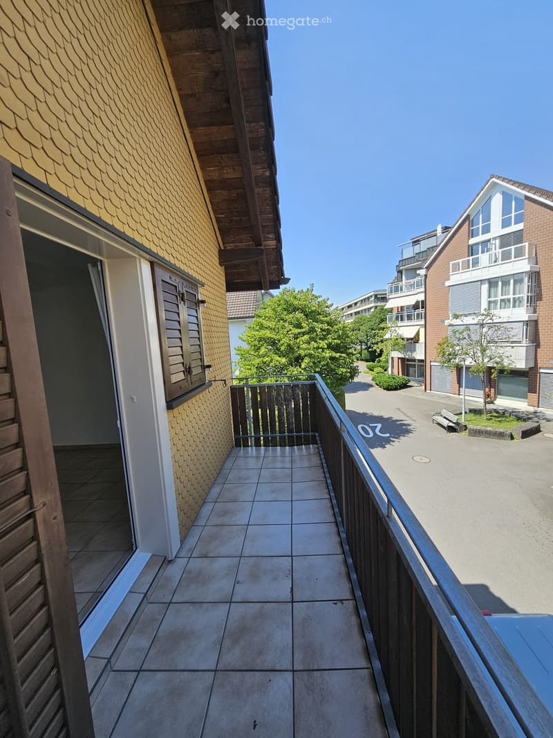 Balcony with tiled floor, metal railing, and view of a residential area with buildings, trees, and a bench.