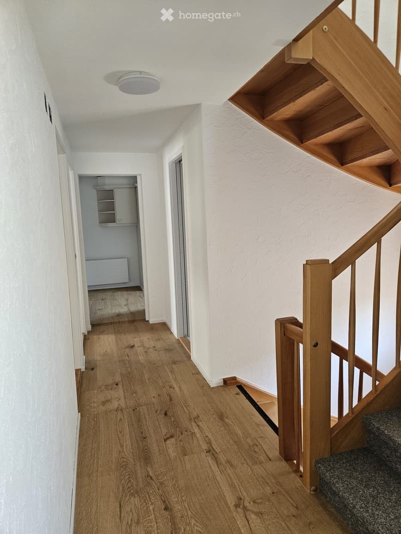 A well-lit hallway with wooden flooring, white walls, and a staircase leading upwards. The staircase features wooden railings and steps, with a small open shelving unit visible in the background.