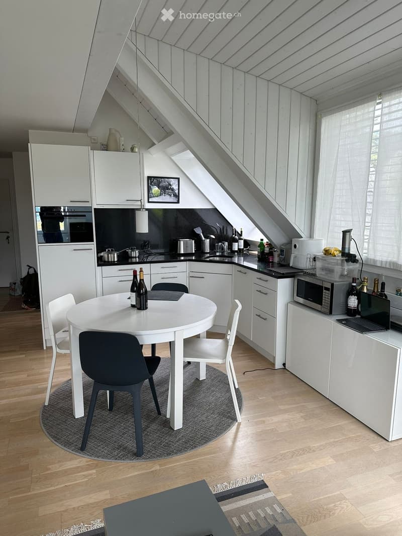 Modern kitchen with white cabinets, a round dining table with chairs, and various appliances including a microwave and oven. The space features wooden flooring and large windows with blinds.