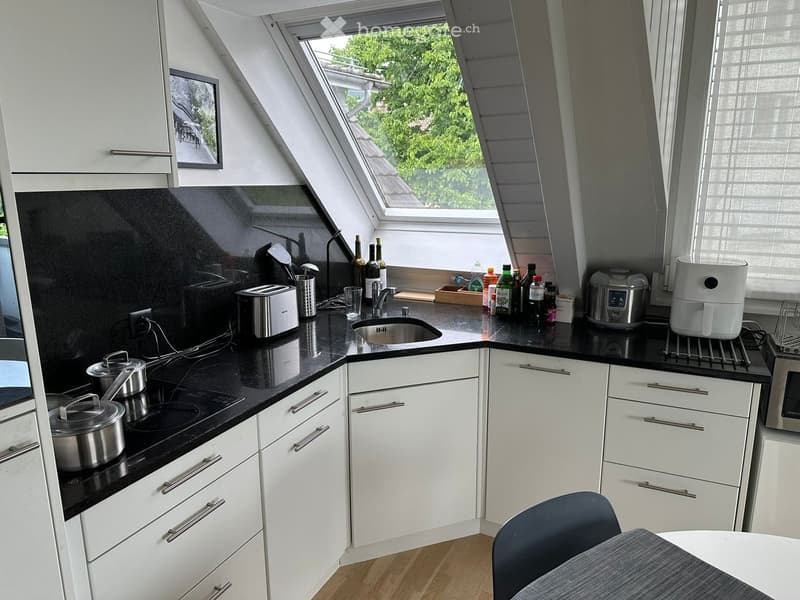 Modern kitchen with white cabinets, black countertops, and stainless steel appliances. Features include a sink, stove, microwave, toaster, and various kitchen utensils. A small dining area with a table and chair is visible.