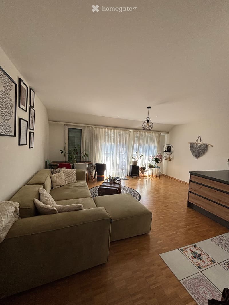 Modern living room with a sectional sofa, coffee table, chairs, and a dining area with a table and chairs. The room features wooden flooring, large windows with curtains, and decorative elements like picture frames and a hanging decor.