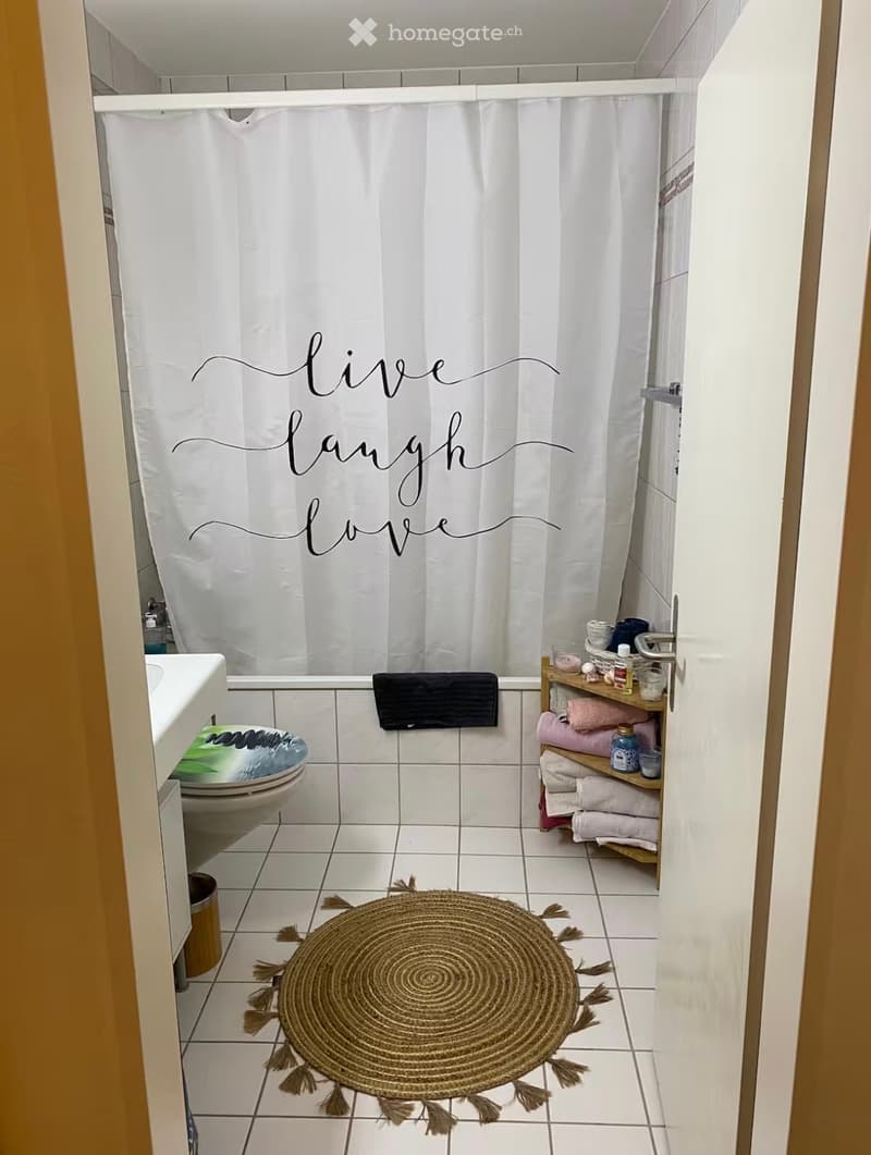 A small bathroom with white tiled walls and floor, a toilet, a sink, a shower curtain with the words "live laugh love" written on it, a towel rack with towels, and a round woven bath mat with tassels.