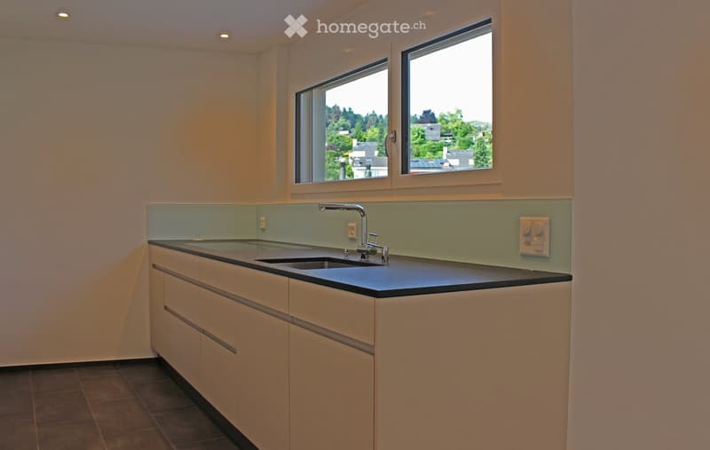 Modern kitchen with light-colored cabinets, black countertop, and a sleek faucet. The backsplash is a light green color, and the room has large windows offering a view of greenery outside.