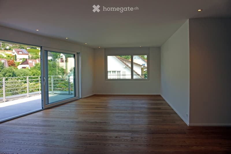 Empty room with wooden flooring, large sliding glass door leading to a balcony, and a window overlooking a residential area.