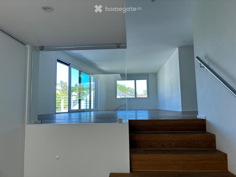 Modern, open-concept hallway with wooden flooring, white walls, and a glass partition leading to a bright room with large windows and a view of greenery.