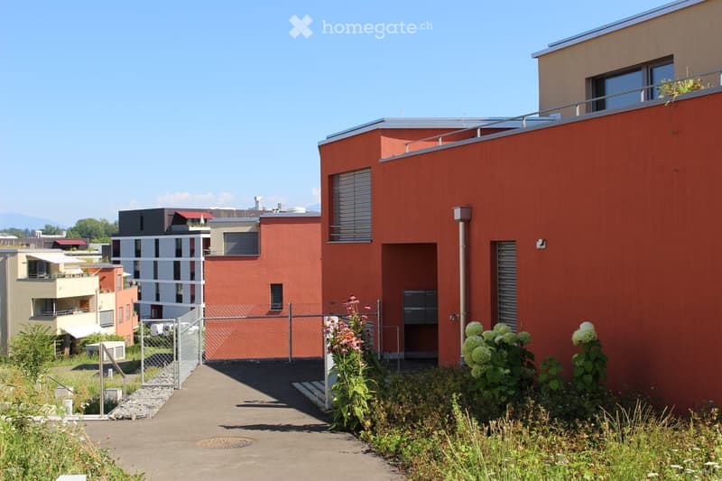 Modern residential building with red facade, multiple windows, and a small garden area in front.