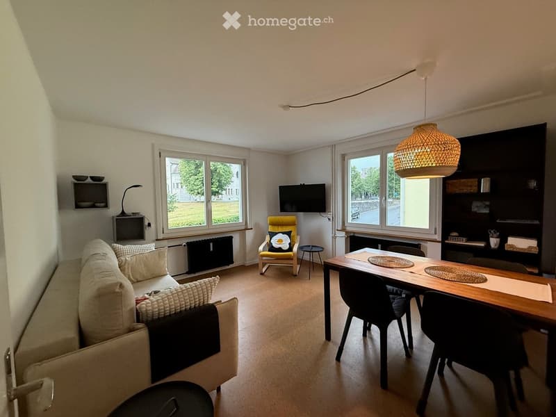 Bright living and dining area with large windows, a cream-colored sofa, a wooden dining table with chairs, a hanging woven lamp, and a television mounted on the wall.