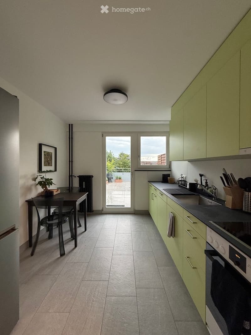 Modern kitchen with light green cabinets, stainless steel appliances, and a dining area with a table and chairs. The space features tiled flooring, a large window, and a door leading to a balcony.