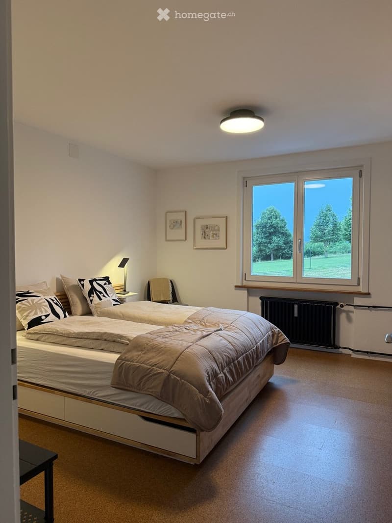Bright bedroom with two single beds, nightstand, lamp, chair, radiator, and framed pictures on the wall. Large window offers a view of greenery.