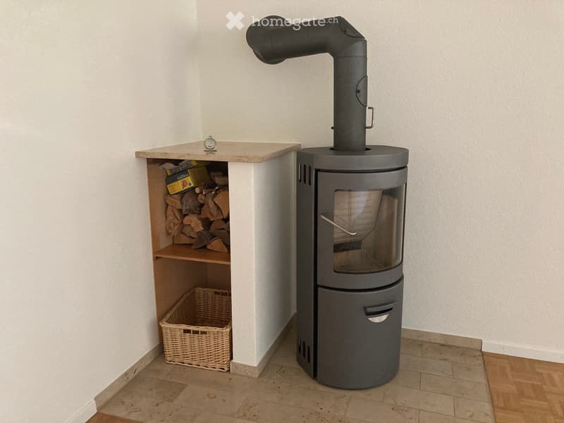 Modern wood-burning stove with a glass door and chimney, accompanied by a wooden shelf holding logs and a wicker basket.