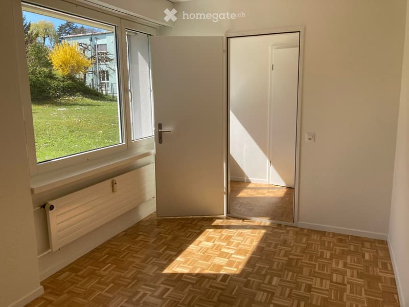 Empty room with wooden flooring, large window with view of garden, white walls, and a closed door.