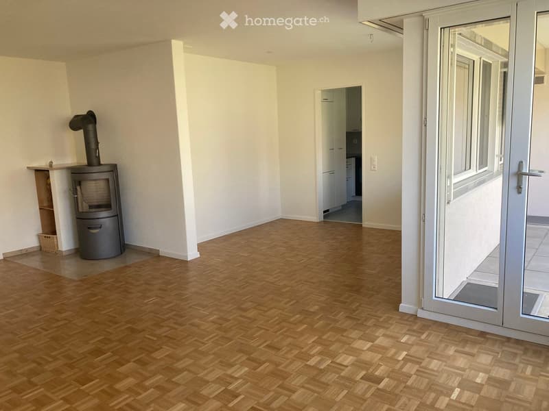Empty hallway with wooden flooring, white walls, and a visible fireplace in the corner. A doorway leads to another room with white cabinets.