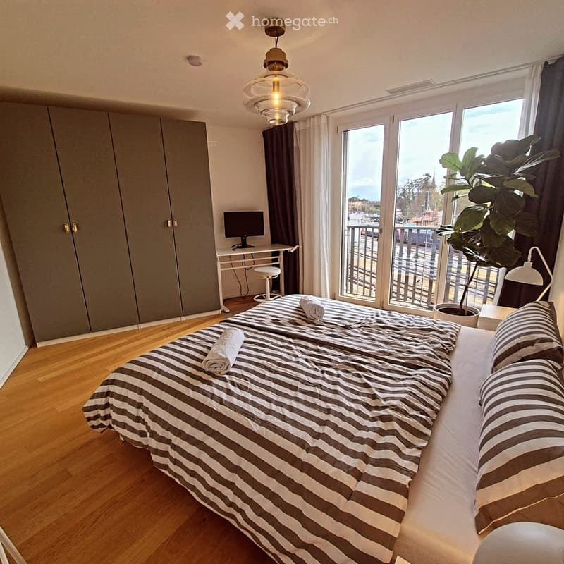 Modern bedroom with a large bed, striped bedding, a desk with a screen, a stool, a lamp, and a potted plant near the balcony.