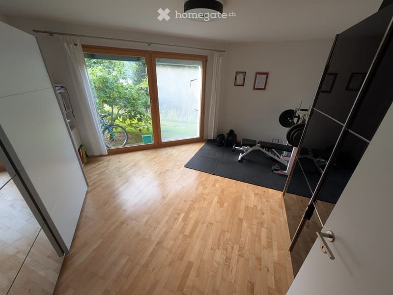 Empty room with wooden flooring, large windows, and a bicycle visible outside. No furniture or equipment present.