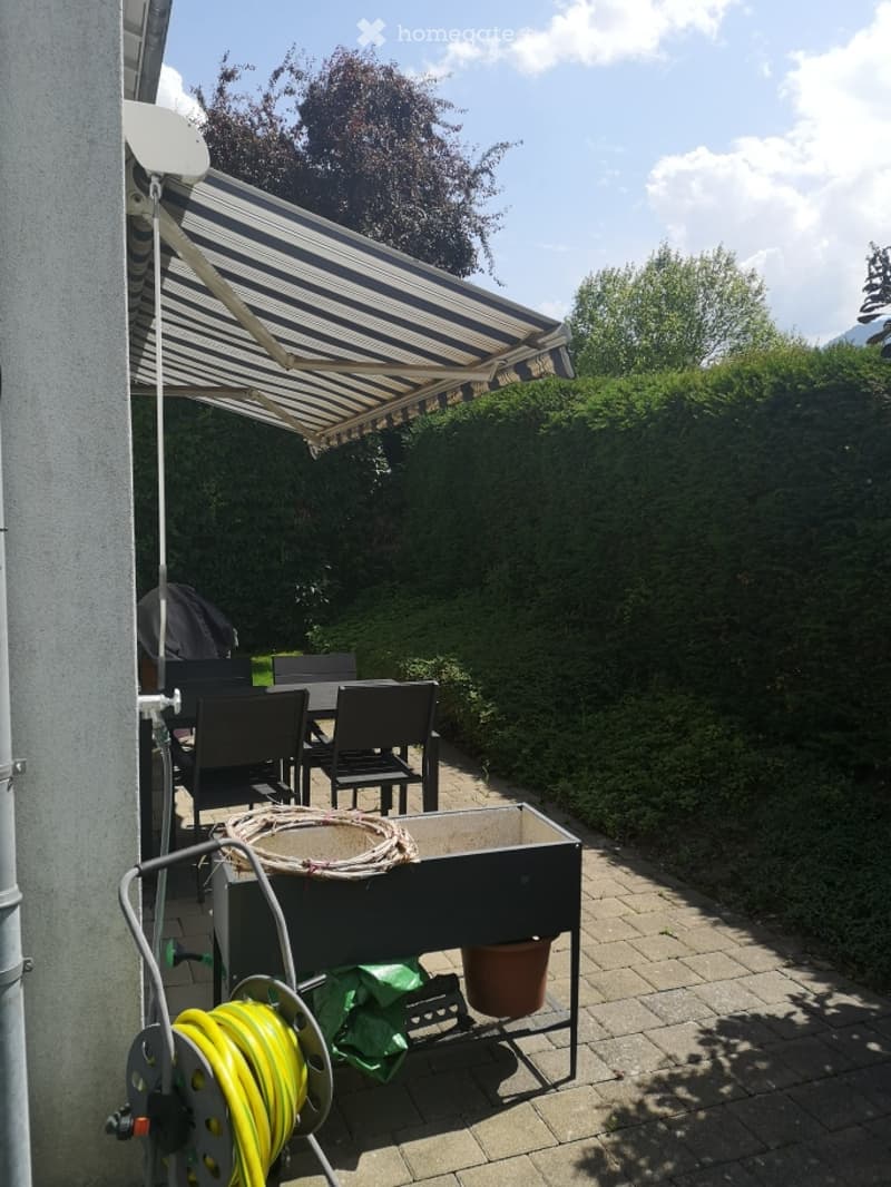 Outdoor patio area with a striped awning, several black chairs and a table, a coiled yellow hose, and a storage box with a coiled rope and a green bag.