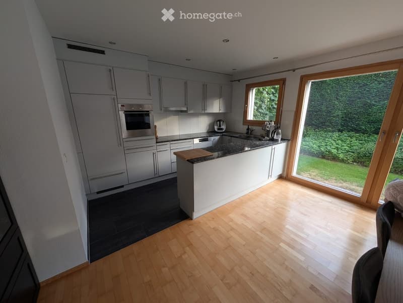 Modern kitchen with light grey cabinets, stainless steel appliances, wooden flooring, and large windows overlooking a green garden.