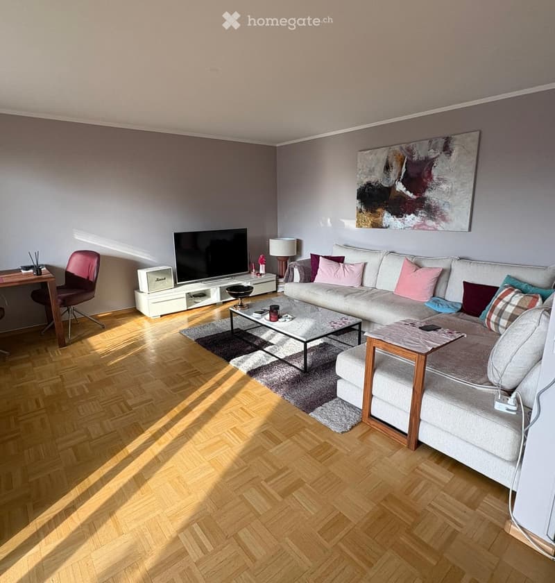 Modern living room with a grey sectional sofa, coffee table, side table, TV, and a wooden desk with a chair. The room features wooden flooring and a large abstract painting on the wall.