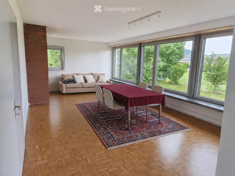 Bright living room with large windows, wooden flooring, a sofa, a dining table with chairs, and a patterned rug.