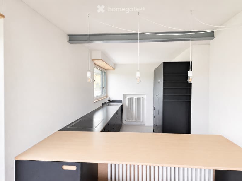 Modern kitchen with white walls, black countertops, and a light wood island. Features pendant lights, a sink, stove, and built-in cabinets.