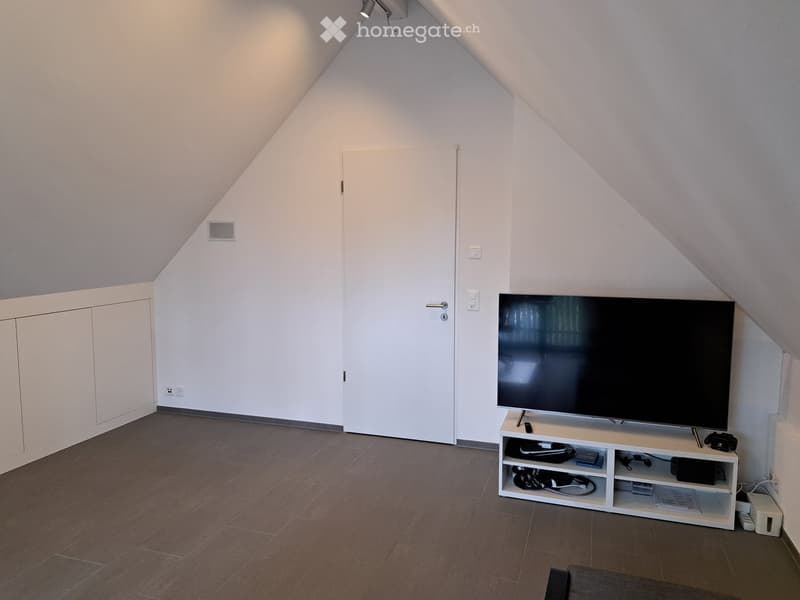 Modern attic room with slanted ceilings, tiled floor, white walls, and a TV on a stand.