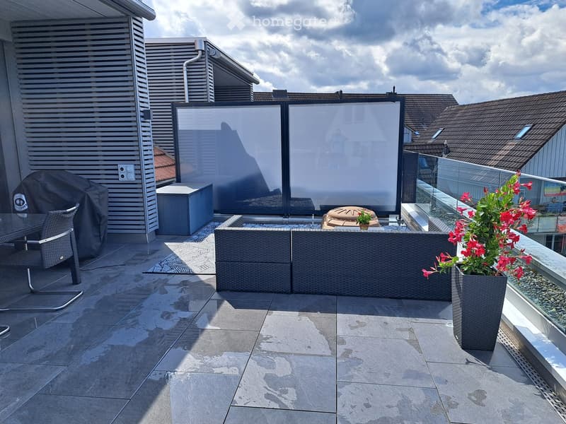 A modern rooftop terrace with a tiled floor, glass railing, and outdoor furniture including a sofa, chairs, a table, and flower pots with red flowers.