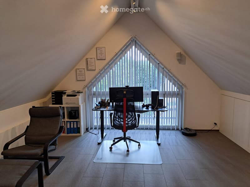 Office space with a desk, chair, screen, laptop, printer, and a comfortable armchair, located in an attic with large windows and slanted ceilings.
