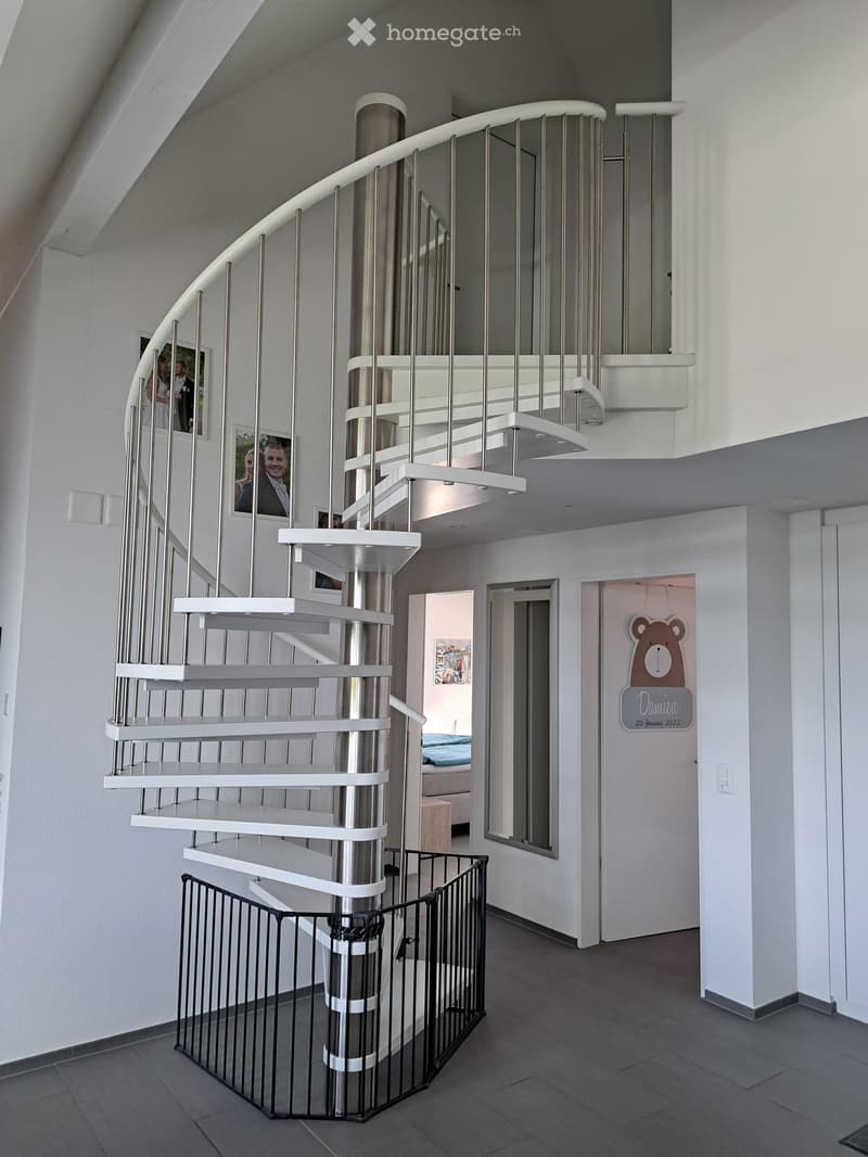 Modern interior hallway featuring a sleek, white spiral staircase with metallic railings, a safety gate at the bottom, and visible doorways leading to other rooms.