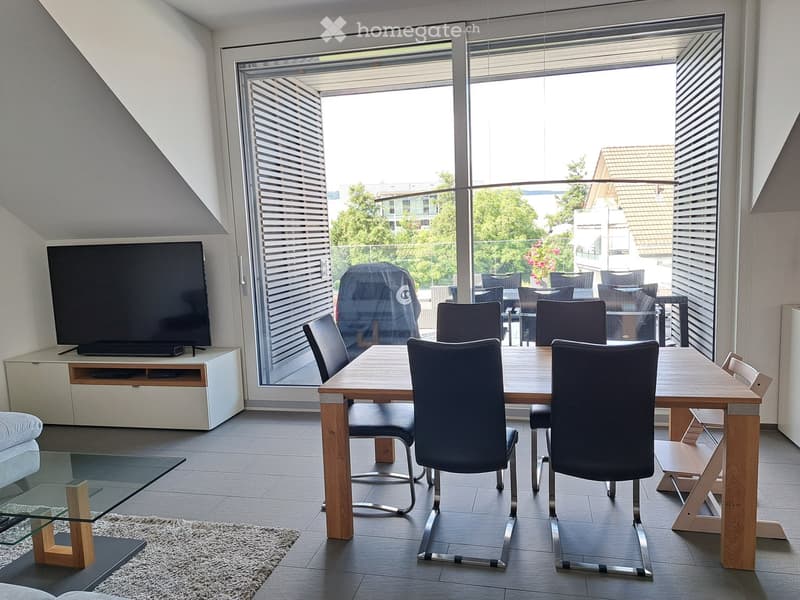 Bright, modern dining and living area with large windows offering a view, featuring a wooden dining table with chairs, a TV on a white stand, and a glass coffee table.