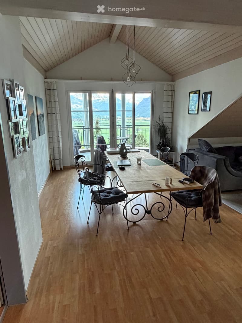 Bright living room with wooden flooring, large windows offering a scenic mountain view, a sofa, a dining table with chairs, and decorative picture frames on the walls.