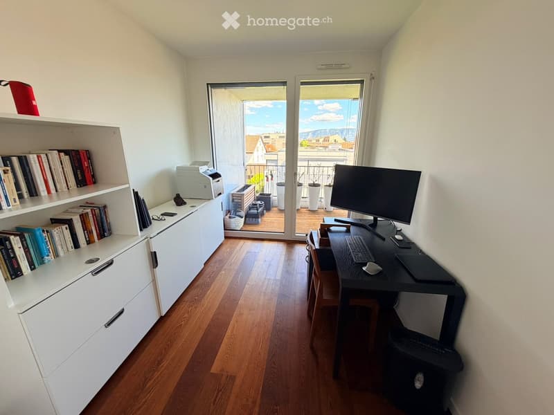 Modern, well-lit room with wooden flooring, a desk setup, a large window opening to a balcony, and built-in white cabinetry with books and a printer.