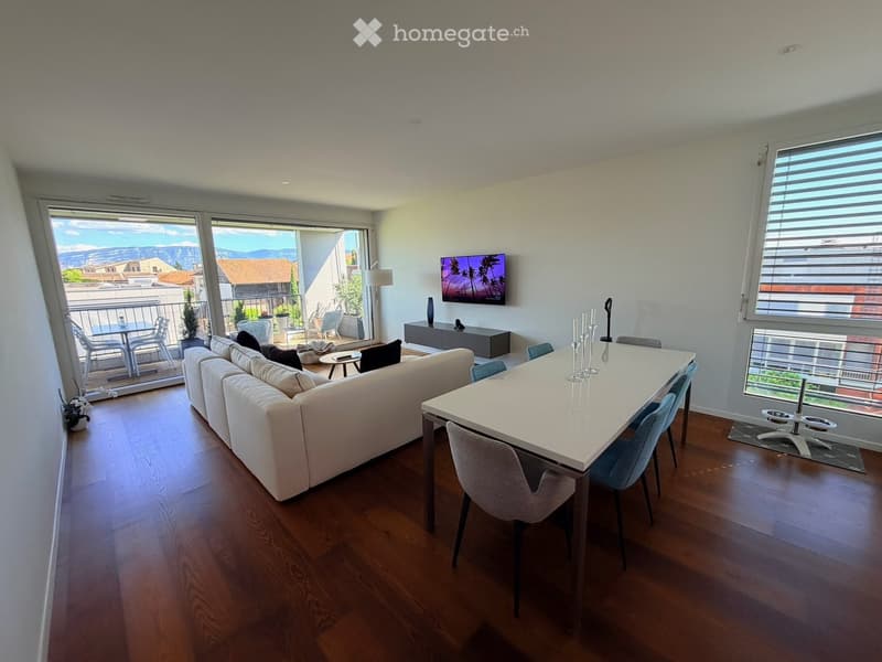 Modern living and dining area with a white sectional sofa, a white dining table with chairs, a television mounted on the wall, and a view of the mountains through large windows and a balcony.